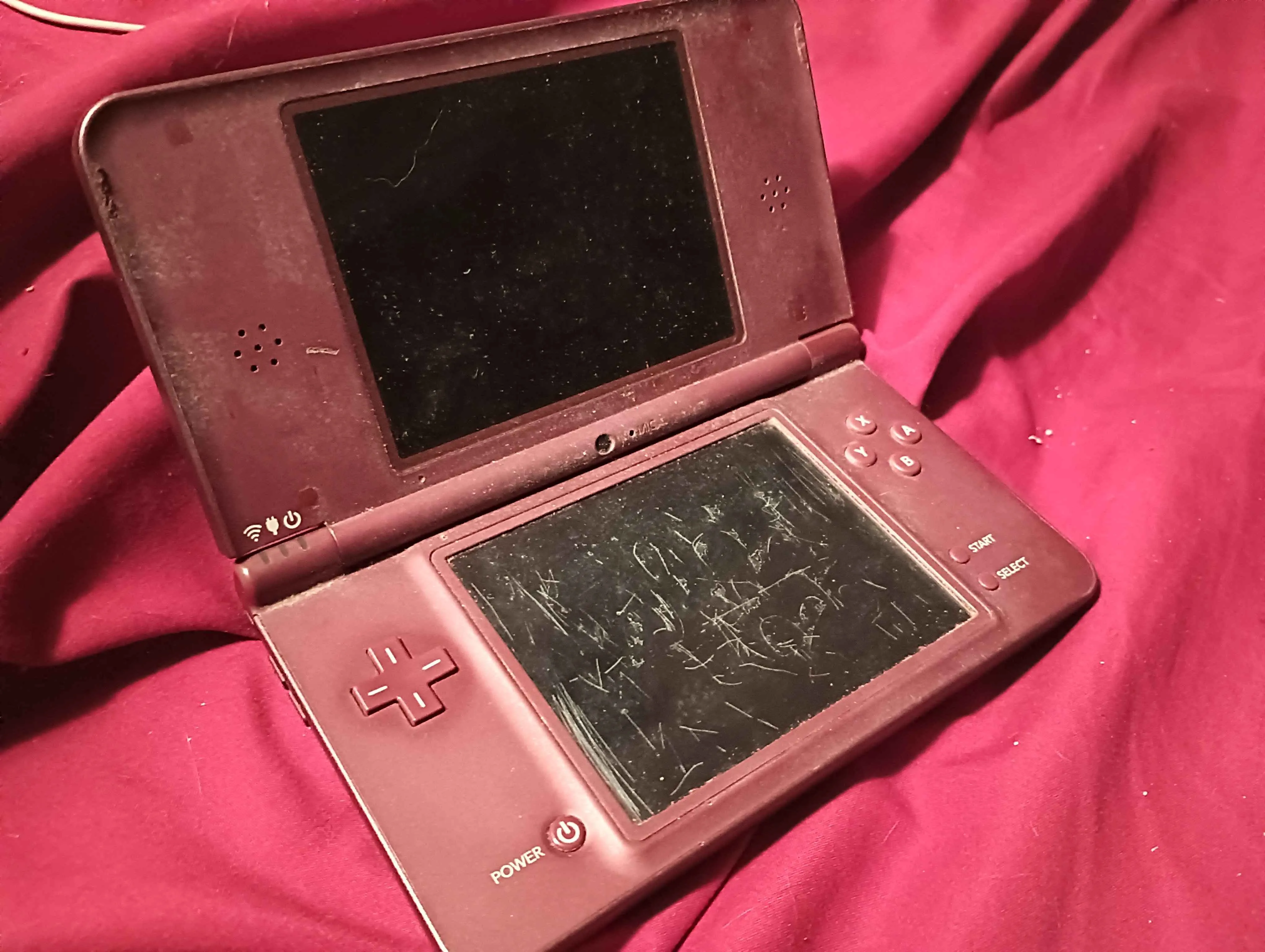 photo of a burgundy dsi, open but turned off. The bottom screen is horribly scratched up and the console is overall worn and dusty.